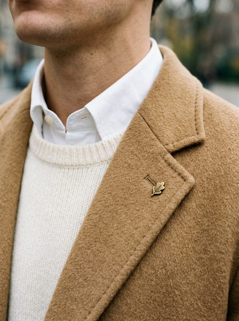 Close-up of a white shirt collar above an ivory knit under a camel coat with a small lapel pin