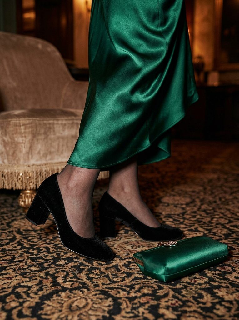 Black velvet block-heel pumps under an emerald satin skirt beside a small satin clutch